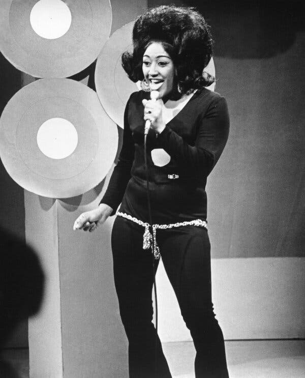 A black-and-white photo of Jean Knight singing on a television show. She wears a dark pantsuit and holds a microphone in her left hand. Her hair is piled high.